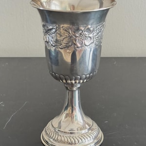 May include: A silver-toned metal goblet featuring a flared rim and a polished finish. The upper portion of the bowl is decorated with an embossed floral pattern and textured detailing, while the base displays a repeating rope-like border design.
