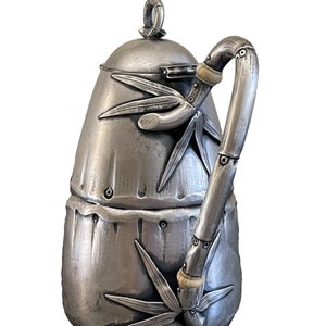 May include: A silver teapot with a bamboo design. The teapot has a round lid and a handle shaped like a bamboo stalk. The teapot is decorated with leaves and a bamboo stalk.