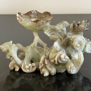 Puede incluir: Una escultura de jade tallada de una flor de loto estilizada con un pequeño plato en la parte superior. La escultura está intrincadamente detallada y tiene un color verde claro con acentos marrones.