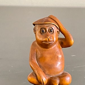 May include: A small, wooden monkey figurine with a hat, sitting with one hand on its head. The monkey has brown eyes and a brown body.