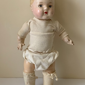 May include: A vintage porcelain doll with blonde hair, blue eyes, and rosy cheeks. The doll is wearing a white shirt, white diaper, and white knitted booties.