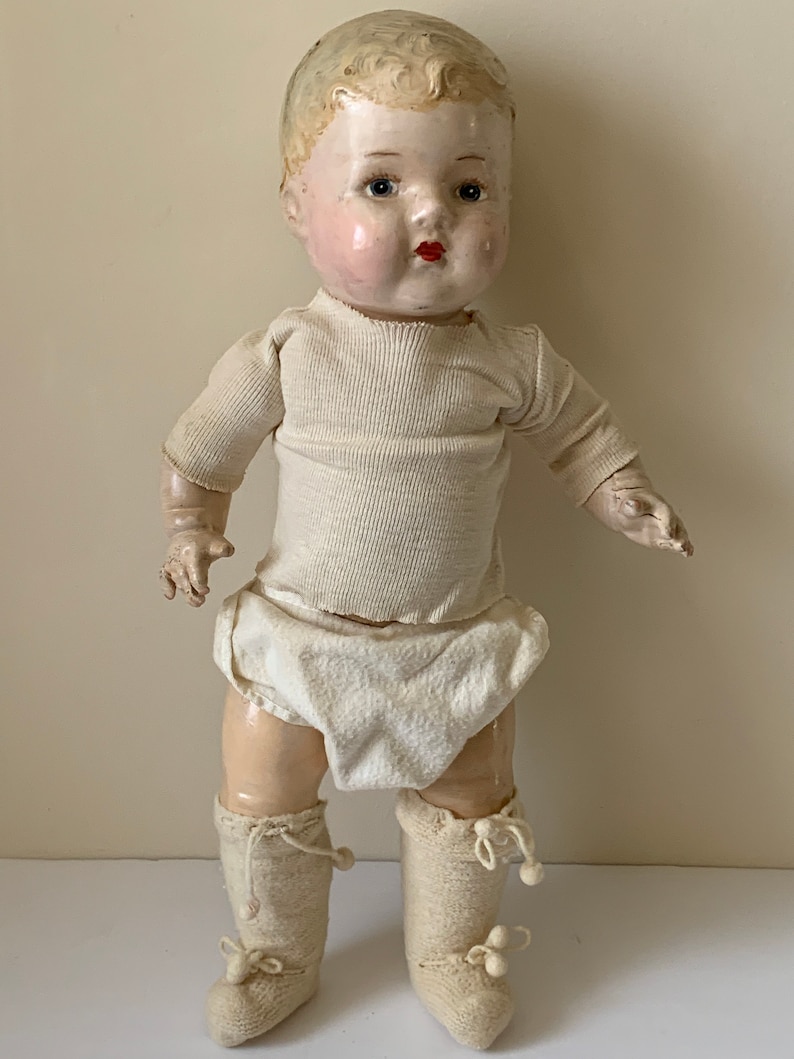May include: A vintage porcelain doll with blonde hair, blue eyes, and rosy cheeks. The doll is wearing a white shirt, white diaper, and white knitted booties.