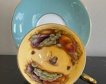 May include: A yellow teacup with a gold rim and a fruit design. The cup is sitting on a light blue saucer with a gold rim.