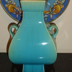 Pode incluir: Um vaso de cer&acirc;mica turquesa com duas al&ccedil;as e uma base quadrada. O vaso tem um acabamento brilhante e &eacute; decorado com um efeito degrad&ecirc; sutil.