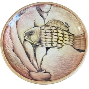 May include: A round, ceramic plate with a hand-drawn illustration of a fish. The fish is tan and yellow, with a detailed scale pattern. The plate has a light brown rim and the name "MANUEL" is written on the side.