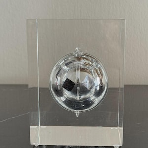 May include: A clear glass sculpture featuring a spherical element encased within a rectangular block. The sphere has internal details and a small, dark square. The sculpture is displayed on a dark surface.