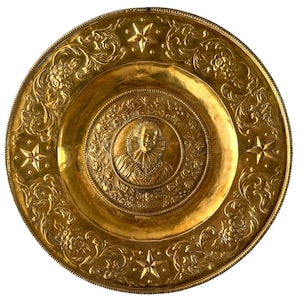 Puede incluir: Un plato decorativo redondo de color dorado con intrincados patrones florales y de estrellas en relieve. El centro presenta un retrato de una persona con un cuello de lechuga. El plato tiene una superficie brillante y reflectante.