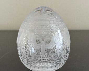 Könnte beinhalten: Ein klares, eiförmiges Dekorationsobjekt mit aufwendigen geätzten Designs. Das Design umfasst einen doppelköpfigen Adler, Lorbeerkränze und florale Muster. Das Objekt besteht aus Glas und steht auf einer dunklen Oberfläche.
