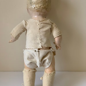 May include: A vintage porcelain doll wearing a white knit onesie, diaper, and knitted booties. The doll has a painted face and blonde hair.