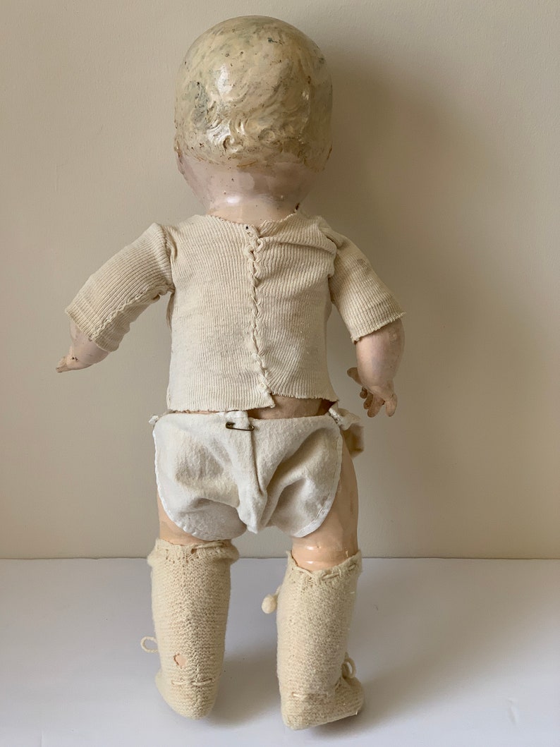 May include: A vintage porcelain doll wearing a white knit onesie, diaper, and knitted booties. The doll has a painted face and blonde hair.