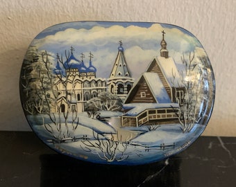 May include: A hand-painted wooden box with a winter scene of a church and houses covered in snow. The box is oval shaped and has a blue and white background.