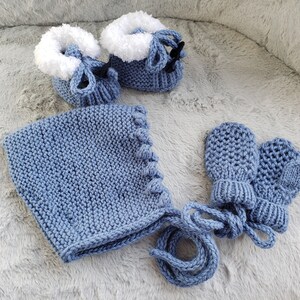 Può includere: Un berretto di lana blu per neonato con un set abbinato di guanti e scarpine. Il berretto ha un laccio sul retro e le scarpine hanno un bordo bianco.