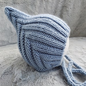 Könnte beinhalten: Eine hellblaue gestrickte Baby-Mütze mit einer langen, spitzen Spitze und einer passenden Schleife. Die Mütze ist mit einer Rippenstrick-Masche gearbeitet und hat ein weiches, gemütliches Gefühl.