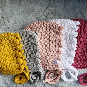 May include: Four knitted baby bonnets in different colors: yellow, gray, pink, and red. Each bonnet has a row of small pom-poms around the edge and ties at the back. The bonnets are laid out on a gray fuzzy surface. The pink bonnet has a tag that says "Liddy's".