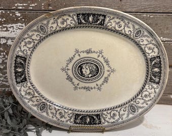 May include: Oval serving platter with a cream-colored surface and black decorative border. The border features floral and geometric patterns, with a central medallion depicting a classical profile. The platter is supported by a gold-colored metal stand.