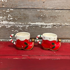 May include: Two ceramic Christmas mugs shaped like red boots with white trim and holly berry accents. The handles are striped in red, white, and green. Each mug has a gold-colored ball on the toe.