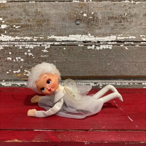 May include: A vintage angel doll with a white dress, wings, and fluffy hair, lying on a red surface. The doll has a painted face with blue eyes and rosy cheeks. The angel is a decorative item.