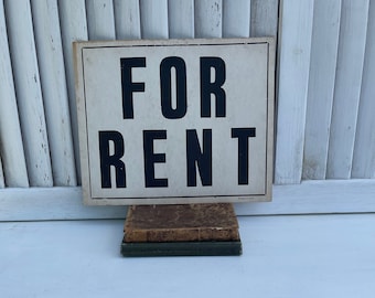 Peut inclure: Une enseigne vintage "FOR RENT" avec des lettres noires sur fond crème, posée sur deux livres anciens. L'enseigne est imprimée aux États-Unis et est placée devant un fond de volets blancs.