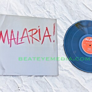 Könnte beinhalten: Eine Schallplatte mit einem schwarzen Label und einem roten Label mit dem Text "MALARIA!" darauf. Die Schallplatte liegt auf einer weißen Oberfläche mit dem aufgedruckten Text "BEATEYEMEDIA.COM".