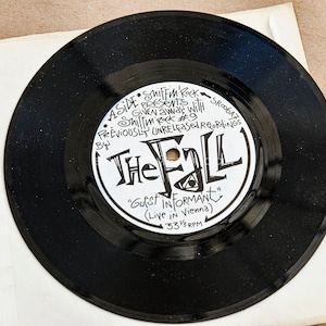 Op de afbeelding: Een zwarte vinylplaat met een wit label. Het label heeft de tekst "A Side Shiffm Rock Presents Sroogaz Given away with Smillm Rock #9 Previously Unreleased Recordings By The Fall "Guest Informant" (Live in Vienna) 33 1/3 RPM".