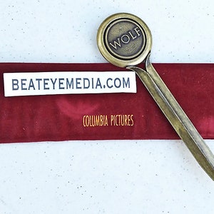 May include: A gold-colored letter opener with a round handle featuring the word "WOLF" in raised letters. The handle is attached to a long, pointed blade. The letter opener is resting on a red velvet case with the text "BEATEYEMEDIA.COM" and "COLUMBIA PICTURES" printed on it.