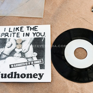 Könnte beinhalten: Eine schwarz-weiße 7-Zoll-Vinyl-Schallplatte mit einem weißen Label, auf dem "I Like the Sprite in You." steht. Die Schallplatte liegt auf einer braunen Oberfläche. Der Text "The Blockbuster Hit of the Summer!" und "Mudhoney" ist auf dem Plattencover gedruckt.