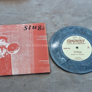 Op de afbeelding: Een zwart-witte 7-inch vinylplaat met een grijs label. Het label heeft de tekst "Sympathy for the Second Industry" en "Slug: Breathe The Thing Out".