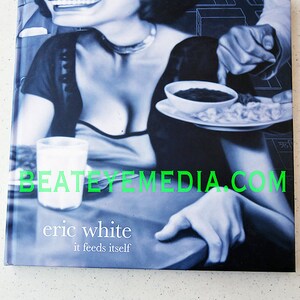 May include: A black and white photo of a woman with a plate of food in front of her. The text "eric white it feeds itself" is printed on the bottom of the photo. The website "beateymedia.com" is printed in green letters across the bottom of the photo.
