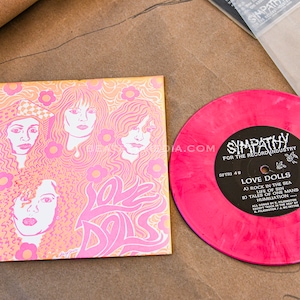May include: A pink vinyl record and album art for "Love Dolls" by Sympathy for the Record Industry. The album art features a pink and orange floral design with illustrations of women. The record label is black with white text.