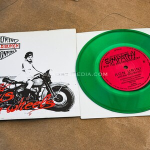 Op de afbeelding: Een vintage vinylplaat en hoes. De hoes heeft een zwart-wit illustratie van een persoon op een motorfiets met de tekst "World Venus on Wheels". De groene plaat heeft een rood label met de tekst "Sympathy".