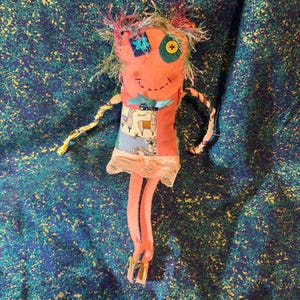 May include: A handmade rag doll with pink fabric, a colorful patchwork dress, and a whimsical face with one green button eye and one blue flower eye.