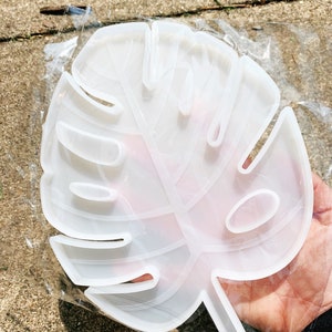 Puede incluir: Un molde transparente en forma de hoja, hecho de plástico flexible. El molde está en una bolsa de plástico transparente. El diseño de la hoja tiene varias secciones recortadas y un tallo. El molde se utiliza probablemente para proyectos de arte con resina o artesanía.