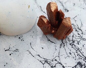 May include: A small, copper-colored crystal cluster with a glittery finish. The crystal is sitting on a white surface with black splatter paint.
