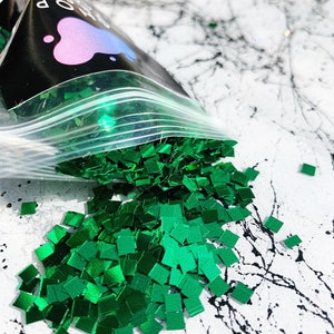Op de afbeelding: Een doorzichtige plastic zak gevuld met groene vierkante glitter, die uitloopt op een wit en zwart marmer oppervlak.
