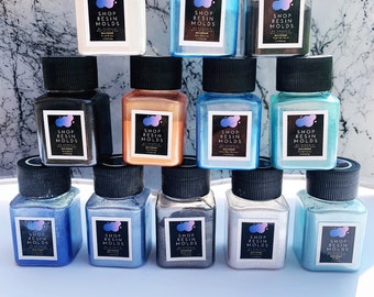 May include: Twelve small bottles of resin pigment in various colors, including white, blue, black, brown, gold, and silver. Each bottle has a black lid and a label that reads "Shop Resin Molds".