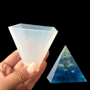 Può includere: Uno stampo in silicone trasparente a forma di piramide, utilizzato per la colata di resina. Lo stampo è tenuto in una mano, e sullo sfondo è visibile una piramide in resina finita con un motivo floreale blu e bianco.