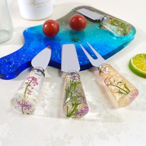 Puede incluir: Un juego de tabla de quesos y cuchillos para queso. La tabla es azul y verde con un diseño de galaxia. Los cuchillos tienen mangos de resina transparente con flores secas. Dos tomates cherry rojos están en la tabla.