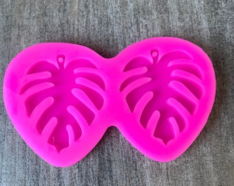 May include: Pink silicone mold with two cavities shaped like monstera leaves. The mold is designed for making earrings or other small crafts.