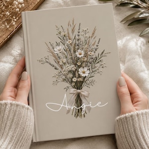 Puede incluir: Un diario de tapa dura en color beige claro con una ilustración impresa de un ramo de flores silvestres secas atado con una cinta. El nombre Angie está escrito en letra cursiva blanca debajo del arreglo floral en la portada del libro.