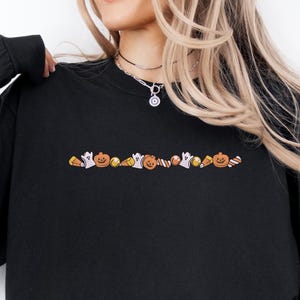 May include: Black long-sleeve shirt with an embroidered Halloween-themed design across the chest. The design includes pumpkins, ghosts, and candy corn. The shirt is paired with blue jeans.