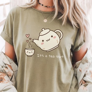 Pode incluir: Camiseta verde-oliva com um desenho animado de uma chaleira despejando chá em uma xícara, com corações flutuando. O texto "It's a tea shirt" está impresso abaixo. A camiseta tem mangas enroladas.