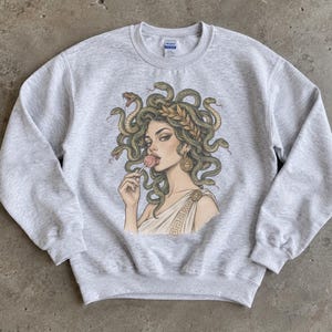 Peut inclure: Un sweat-shirt à col rond gris clair orné d'une illustration graphique représentant une figure aux cheveux en forme de serpents, portant une couronne de laurier et un vêtement drapé, tenant une sucette rose. Le motif inclut des boucles d'oreilles dorées.