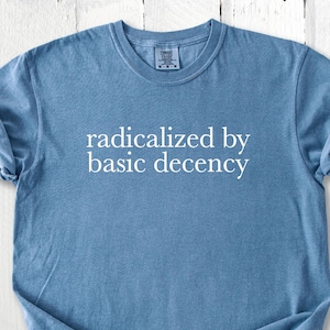 May include: Blue jean-colored t-shirt with the phrase "radicalized by basic decency" printed in white. The shirt has a crew neck and short sleeves, with the sleeves rolled up. The shirt is made of a soft, comfortable material.