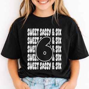 Puede incluir: Camiseta negra con el texto "SWEET SASSY & SIX" en un diseño apilado. El número "6" se muestra de forma destacada en el centro. La camiseta la lleva una persona.