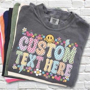 May include: A stack of Comfort Colors t-shirts in various colors, including pink, orange, navy, black, and olive green. The top shirt is charcoal gray with a colorful graphic that says "CUSTOM TEXT HERE" with a smiley face and flowers.