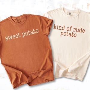 Op de afbeelding: Twee t-shirts, één in een warme oranje kleur en de andere in ivoor. Het oranje shirt heeft de tekst "sweet potato" in een lichtbeige lettertype. Het ivoorkleurige shirt heeft de tekst "kind of rude potato" in een bruin lettertype.