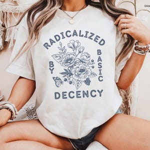 May include: A Gildan white t-shirt featuring the text "RADICALIZED BY BASIC DECENCY" in a circular design with floral and butterfly illustrations in blue. The shirt is a casual wear item.