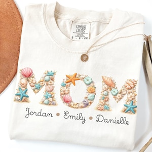 Peut inclure: T-shirt crème avec le mot "MOM" formé de coquillages et d'étoiles de mer. Les noms "Jordan", "Emily" et "Danielle" sont imprimés en dessous. Un collier doré et un chapeau marron sont également visibles.