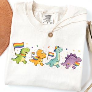 Pode incluir: Uma t-shirt cor de marfim com um gráfico de quatro dinossauros de desenho animado coloridos a caminhar em fila. Os dinossauros são verdes, cor de laranja, azuis e roxos, transportam bandeiras arco-íris e acessórios arco-íris, com corações.