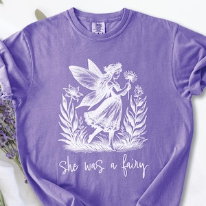 Può includere: T-shirt viola con un'illustrazione bianca di una fata che tiene un fiore, circondata da elementi floreali. Il testo "She was a fairy" è scritto sotto. La maglietta ha le maniche arrotolate.